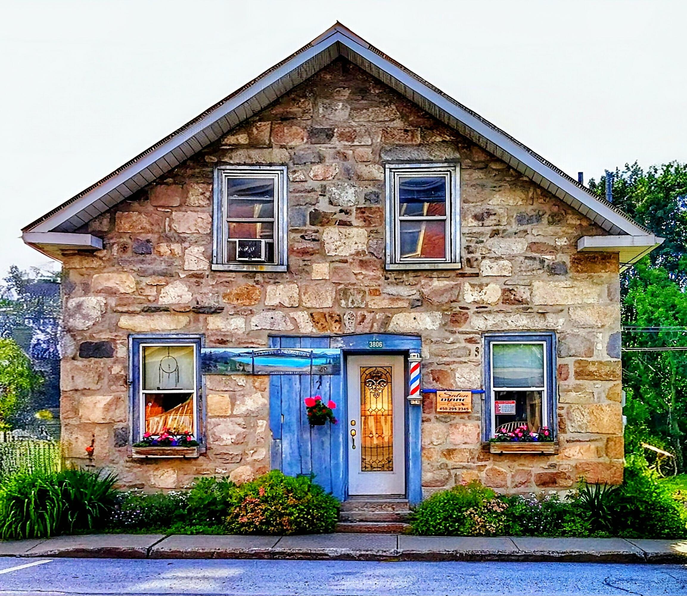 A two-story stone building with a steeply pitched roof is shown, appearing to be a storefront or small house.

The exterior walls are constructed entirely of irregularly shaped, tan and brown stones.
There are four windows visible, two on each floor, each with white frames.

A bright blue wooden door is centered on the ground floor, flanked by windows on either side, and a small sign hangs above it.
The number "306" is visible above the door. 

The windows feature curtains and various objects displayed inside, suggesting a lived-in or business space.
Small flower boxes are attached beneath some of the windows. The building is situated on a paved road with landscaping visible on the sides.
A sign in the right window reads "Barber Styling" in white lettering. 