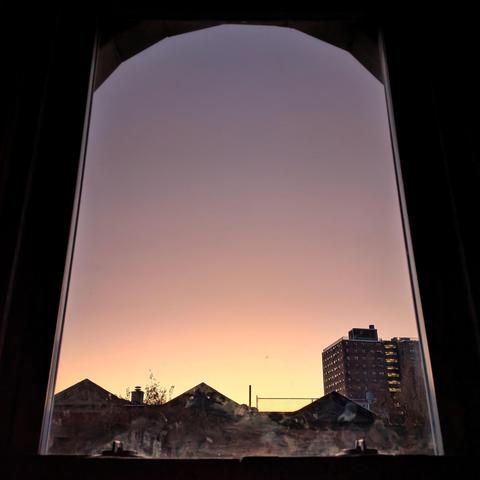Looking through an arched window fifteen minutes before sunrise the cloudless sky fades from lemon yellow at the horizon, through pink,  to dusty gray-blue up above. Pointed roofs of Harlem brownstones are silhouetted across the street, and a taller apartment building can be seen in the distance. 