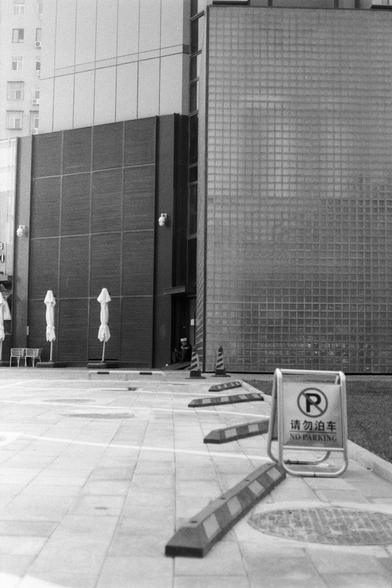 Fujifilm NEOPAN SS (FF)

English Alt Text:
A black-and-white photo of an urban street scene in front of a modern building with glass blocks and metal panels. Several umbrellas and a bench sit near the facade. A clear sign in English and Chinese reads “NO PARKING” (请勿泊车), with cones and barriers enforcing the rule. The area is clean and orderly.

中文替代文字：
这是一张黑白城市街景照片，背景是现代建筑，带有玻璃砖墙和金属外立面。建筑前有几把遮阳伞和长椅。前景中有一个醒目的标志，写着“NO PARKING”（请勿泊车），地面设有交通锥和栏杆以禁止停车。整体环境整洁有序。