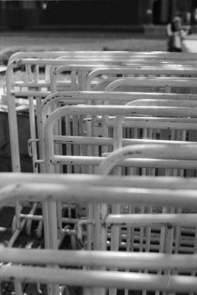 Fujifilm NEOPAN SS (FF)

English Alt Text:
A close-up black-and-white photo of several metal railings, possibly from a hospital or institution, arranged in rows. The frames are worn, with chipped paint and scratches. In the blurred background, human figures and architectural elements suggest an outdoor setting. The repetition of shapes creates a stark, industrial mood.

中文替代文字：
这是一张黑白特写照片，展示了多张金属支架，可能来自医院或机构，整齐排列。床架表面有划痕和掉漆，显得陈旧。背景模糊中可见人影与建筑结构，暗示为户外场景。重复的几何形状营造出冷峻、工业化的氛围。
