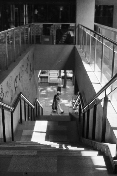 Fujifilm NEOPAN SS (FF)

English Alt Text:
A monochrome image looking down a modern stairwell with metal railings and glass panels. Sunlight casts sharp shadows across the steps. On the left wall, graffiti adds an urban touch. At the bottom, two people walk away, while another figure stands in the distance. The composition emphasizes geometry, light, and human presence.

中文替代文字：
这是一张黑白照片，从楼梯顶端俯视现代楼梯间。金属栏杆与玻璃围栏构成简洁线条，阳光在台阶上投下鲜明阴影。左侧墙面有涂鸦，增添都市气息。楼梯底部有两人行走，远处还有一人站立。画面突出几何结构、光影变化与人的存在感。