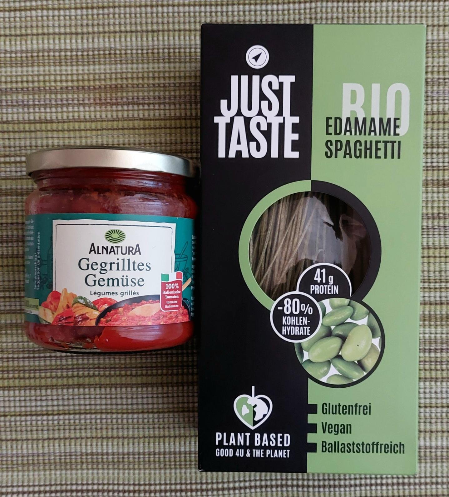 Ein Glas Alnatura Gegrilltes Gemüse-Tomatensoße und eine Packung JustTaste Edamame-Spaghetti liegen auf einem gestreiften moosgrünen Platzdeckchen.