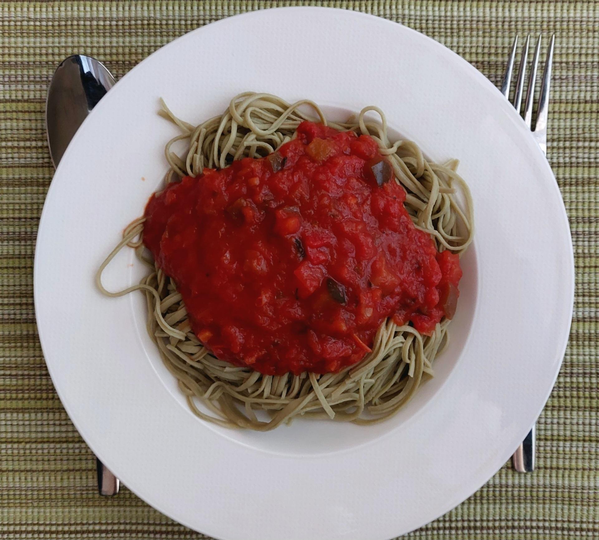 Grüne Spaghetti mit roter Tomatensauce mit Gemüsestückchen auf einem weißen Teller, behleiter von Gabel und Löffel auf einem gestreiften, moosgrünen Platzdeckchen.