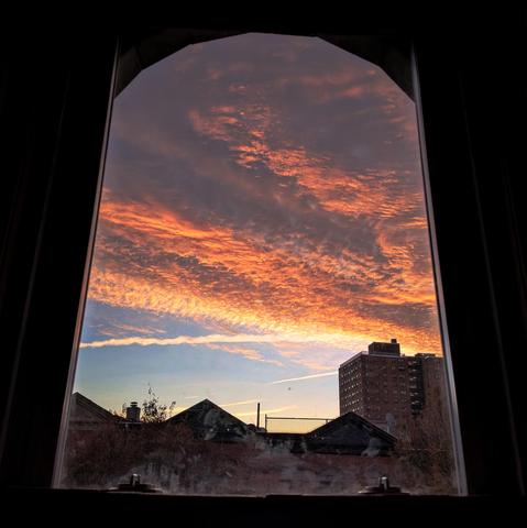 Looking through an arched window three minutes after sunrise there is pale blue sky marked with jet trails at the horizon, while the upper sky is a dense mass or gray and orange finely patterned clouds. Pointed roofs of Harlem brownstones are silhouetted across the street, and a taller apartment building can be seen in the distance. 