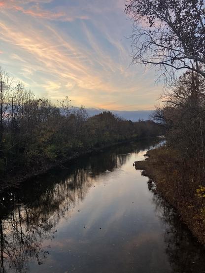 A serene river scene at sunset, featuring soft pastel-colored skies and the reflection of trees along the water's edge. The surrounding landscape includes autumn foliage.