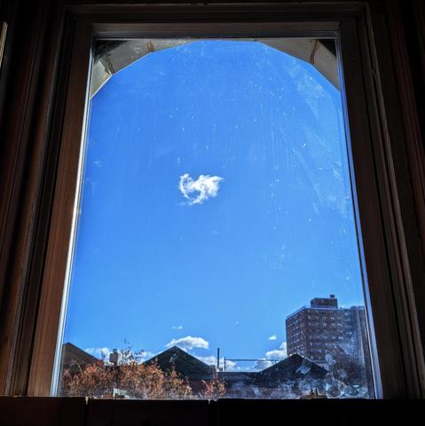 Looking through an arched window two and a half hours after sunrise the deep blue sky has a single, small, puffy white cloud floating in the center. A few other puffy white clouds linger in the distance above the horizon. Pointed roofs of Harlem brownstones are silhouetted across the street, and a taller apartment building can be seen in the distance. The brown tops of two trees are on the bottom and right. The window is grimy.