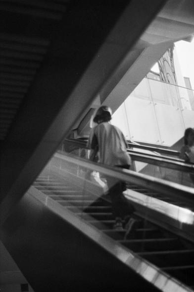 Fujifilm NEOPAN SS (FF)

English Alt Text: A black-and-white photo captures a person ascending an escalator in a modern architectural space. The person wears a hat and casual clothes, seen from a low angle that emphasizes the steep diagonal of the escalator. The surrounding structure includes glass panels and metal beams, forming strong geometric lines. Light and shadow interplay across the surfaces, adding depth and motion. Another person is partially visible on the right, cropped by the frame…