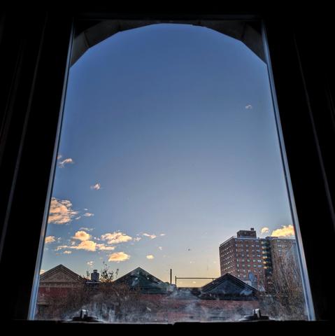 Looking through an arched window forty minutes after sunrise there are a few scattered white puffy clouds in the blue sky above the horizon. Pointed roofs of Harlem brownstones are silhouetted across the street, and a taller apartment building can be seen in the distance. The window is grimy on the bottom.