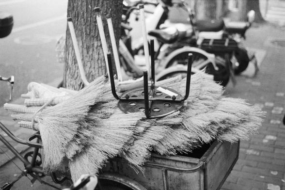 Fujifilm NEOPAN SS (FF)

English Alt Text: A black-and-white street photo showing a bicycle cart loaded with bundles of straw brooms. On top of the brooms sits an upside-down metal stool. The cart is parked along a tree-lined sidewalk, with several scooters and motorcycles in the background. The image captures a slice of urban street commerce, highlighting traditional cleaning tools and the practical use of bicycles for mobile vending.

中文替代文字：
这是一张黑白街头照片，画面是一辆自行车拖车，上面堆满了成捆的稻草扫帚。扫帚上方放着一张倒置的金属凳子…
