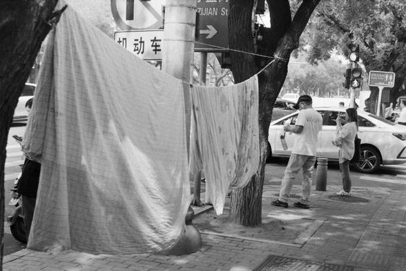 Fujifilm NEOPAN SS (FF)

English Alt Text: A black-and-white photo of a city sidewalk. A man stands near a tree holding a bottle, while a woman stands nearby. Two large cloths hang between trees, possibly drying or acting as barriers. Parked cars line the street, and a traffic light is visible in the background. Signs include “Zijian Street” pointing right, a “No Parking” symbol, and a “Public Toilet” sign with directional arrows. A person wears a shirt with “GUESS” printed on the back. The sce…