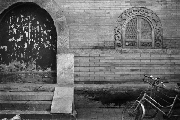 Fujifilm NEOPAN SS (FF)

English Alt Text: A black-and-white photo of an old brick wall with two bicycles leaning against it. On the left is a large arched doorway with a weathered surface and ornate stonework. Next to it is a smaller arched window with intricate carvings and latticework. Below are worn steps and a concrete ramp. The image contrasts aged architectural elements with everyday objects, evoking a sense of history and routine.

中文替代文本： 这是一张黑白照片，展示了一堵老旧砖墙，墙边靠着两辆自行车。左侧是一个大型拱形门洞，表面斑驳，周…