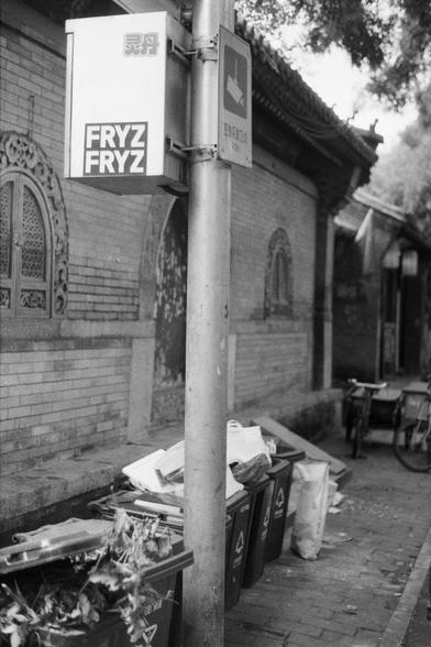 Fujifilm NEOPAN SS (FF)

English Alt Text: A black-and-white image of recycling bins filled with plant debris and bags, placed along a sidewalk beside a brick wall with ornate windows. A pole in the foreground holds two signs: one reads “FRYZ FRYZ” in bold letters, and the other features a broom icon with Chinese characters indicating a sanitation service. Bicycles are parked in the background. The scene juxtaposes modern waste management with traditional architecture.

中文替代文本： 这是一张黑白照片，展示了几排装有…