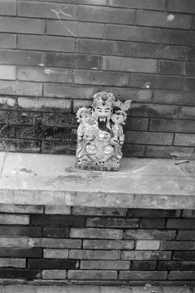 Fujifilm NEOPAN SS (FF)

English Alt Text: A black-and-white photo of a small statue placed on a stone surface against a brick wall. The statue depicts a traditional Chinese figure with a long beard, ornate clothing, and a crown-like headpiece. It holds symbolic objects in its hands. The setting suggests an outdoor or semi-outdoor location, possibly part of a shrine or decorative installation.

中文替代文本： 这是一张黑白照片，展示了一尊放置在石面上的小型雕像，背景是砖墙。雕像描绘的是一位传统中国人物，长须、穿着华丽、头戴冠状饰物，手中持有象征性物品。场景可能是户外或半户外空间，可能属于神龛或…
