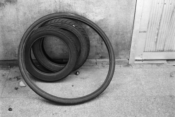Fujifilm NEOPAN SS (FF)

English Alt Text: A black-and-white photo shows three tires leaning against a concrete wall. Two are standard car tires with visible tread, while the third is a thinner bicycle tire placed in front. The setting appears to be a garage or outdoor area, with a tiled floor and a partially visible door on the right.

中文替代文字：
这是一张黑白照片，画面中三个轮胎靠在水泥墙上。两个是普通汽车轮胎，胎纹清晰；另一个是较细的自行车轮胎，放在前方。场景似乎是车库或室外区域，地面铺有瓷砖，右侧有一扇部分可见的门。