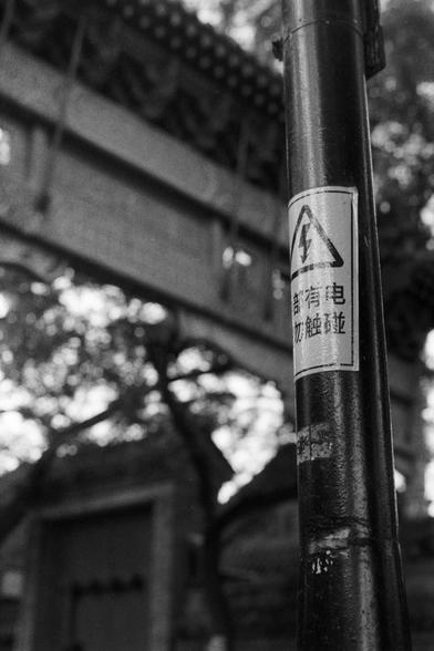 Fujifilm NEOPAN SS (FF)

English Alt Text: A black-and-white photo shows a metal pole with a warning sign attached. The sign features a lightning bolt inside a triangle, indicating high voltage. Beneath the symbol, Chinese text reads “Beware of electricity. Do not touch.” The background includes traditional Chinese architecture, with a tiled roof and ornate gate, suggesting a historical or cultural site. Trees frame the scene, adding a natural element.

中文替代文字：
这是一张黑白照片，画面中一个金属杆上贴有警告标志。标志中是一个三角…