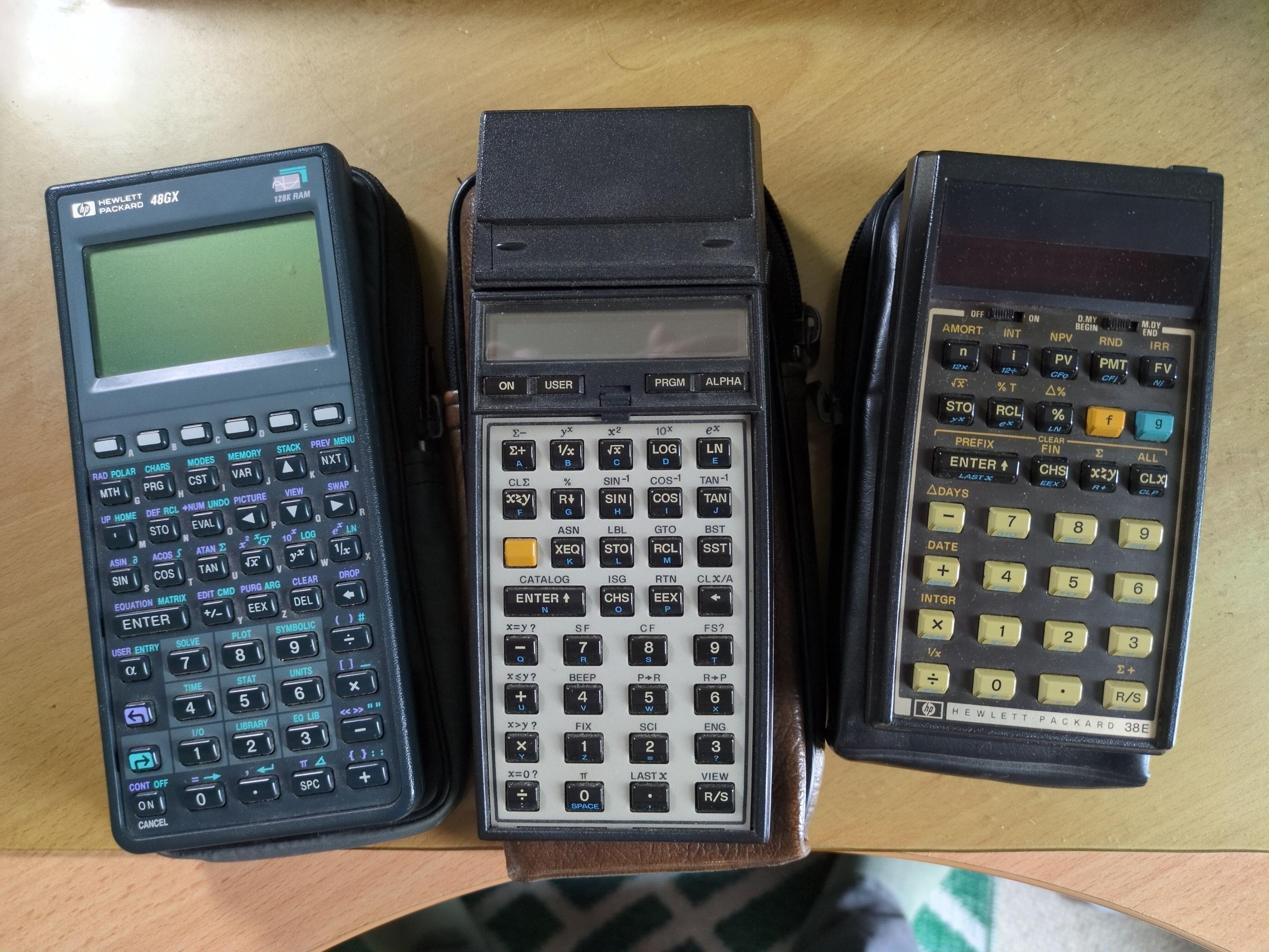 Three old HP calculators, all sitting on their cloth cases. On the left, an HP48GX with a large LCD graphics display. In the middle, an HP 41CV with the add-on magnetic card reader attached. On the right, an HP38E, which has specialised financial functions built in.
