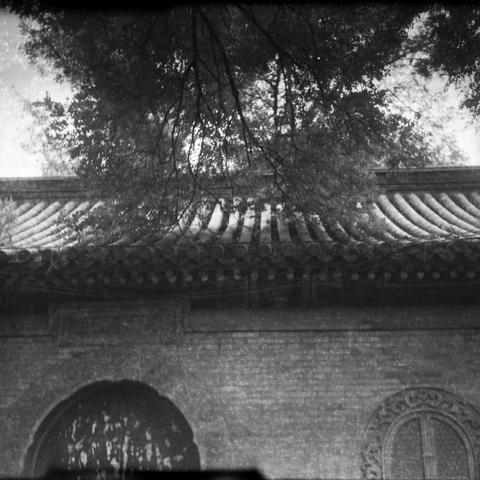 Ilford HP5 Plus 400 (6x6)

English Alt Text
A vintage black-and-white photo of a traditional East Asian building. The roof features cylindrical tiles in overlapping rows, typical of Chinese architecture. Below the roof, a large arched doorway and a circular window with intricate carvings are visible. Tree branches hang in the foreground, partially obscuring the roof. The image has visible grain and wear, suggesting it is archival. The scene is quiet and timeless, evoking cultural heritage and a…