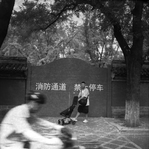 Ilford HP5 Plus 400 (6x6)

English Alt Text
A monochrome photo shows a man standing beside a stroller on a sidewalk. He holds a helmet and faces a wall with Chinese characters reading “消防通道 禁止停车” (Fire Apparatus access route. No parking). The wall is flanked by traditional architectural elements and trees, suggesting a historical urban setting. A cyclist passes by in the foreground, blurred in motion. The contrast between stillness and movement adds depth. The man’s posture is relaxed, and the …