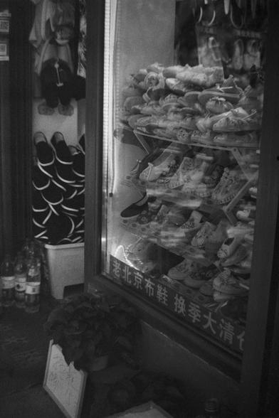 Ilford HP5 Plus 400 (FF)

English Alt Text: A storefront display showcases traditional Chinese cloth shoes arranged neatly behind a glass window. The shoes feature intricate embroidery and colorful patterns, reflecting cultural craftsmanship. To the left, more shoes are stacked in a bin, with hanging items and several bottles on the floor. A potted plant adds greenery near a handwritten sign. The sign and window text read “Old Beijing Cloth Shoes – Seasonal Clearance,” suggesting a local shop o…