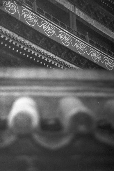 Ilford HP5 Plus 400 (FF)

English Alt Text: A black and white photograph captures the ornate eaves of a traditional East Asian building, possibly a temple or palace. The upper portion features layered wooden beams with swirling cloud motifs painted in white against a dark background. The craftsmanship is intricate, with carved brackets and hanging decorative elements. The lower part of the image fades into soft focus, showing rounded roof tiles typical of classical Chinese or Japanese architect…