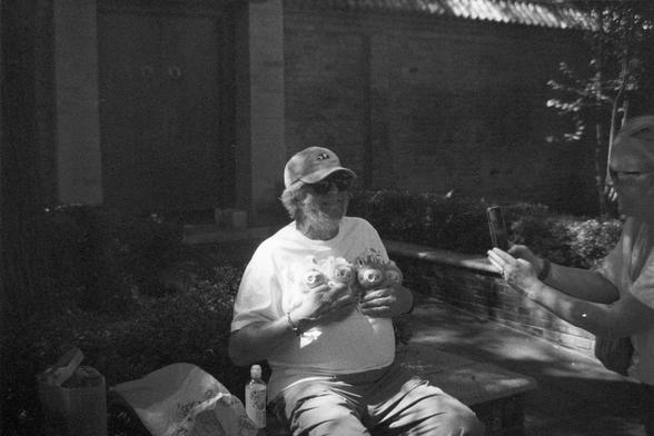 Ilford HP5 Plus 400 (FF)

English Alt Text:
A black-and-white photo captures a candid outdoor moment between two people. One person sits on a bench wearing a cap and sunglasses, holding several plush pig toys in their lap. The other person stands in front of them, holding a smartphone, seemingly taking a photo or video. The background includes leafy trees, bushes, and a brick wall, suggesting a park or garden setting. The mood is playful and lighthearted, with the plush toys adding a whimsical …