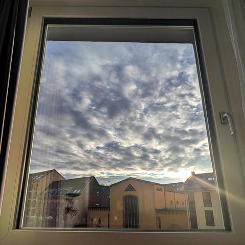 Looking out of a modern window eighty-twi minutes after sunrise, The song is just above the buildings, creating a small lens flare. The sky is full of swelling gray clouds. A few modern buildings are across the street. 