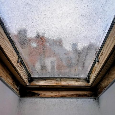 Looking through an attic window ten minutes after sunrise, we can see vague shapes of brick buildings and chimneys, shrouded in light rain. The window has a thick unfinished wood frame. The glass is covered in rain drops and condensation. The sky is blank grey. 