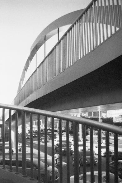 Ilford HP5 Plus 400 (FF)

English Alt Text: A black and white photograph captures an elevated pedestrian bridge spanning a busy urban street. The bridge features vertical railings and an arched support structure above, creating a rhythmic pattern. The perspective is from below, looking up through the railings, which adds depth and layering to the composition. Below the bridge, numerous cars and pedestrians fill the street, suggesting a bustling city environment. The contrast between the rigid g…
