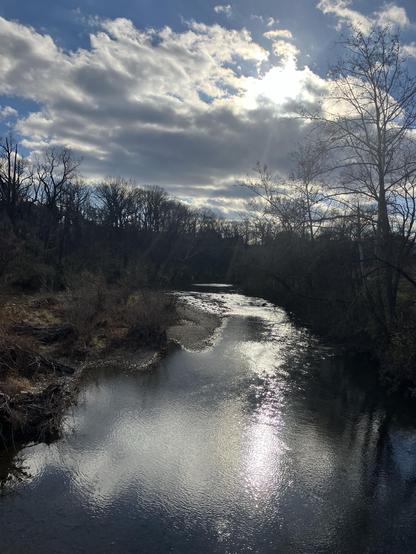 A picturesque river scene with gently flowing water reflecting sunlight. Dense trees line the riverbanks under a backdrop of clouds and a partially visible sun. The atmosphere is serene and natural, showcasing the beauty of the landscape.