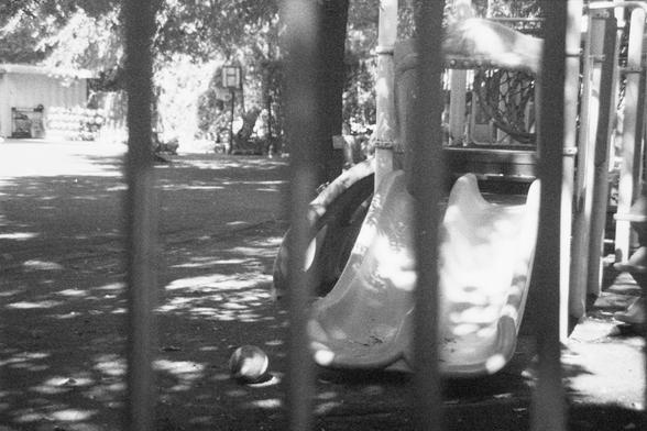 Ilford HP5 Plus 400 (FF)

English Alt Text: A black-and-white image of a playground viewed through vertical bars. In the foreground is a dual-lane plastic slide with a ball nearby. Climbing structures and trees surround the area, casting shadows on the ground. A basketball hoop and a building with shelves are visible in the background. The scene feels quiet and reflective, contrasting playful elements with somber tones.

中文替代文字：
这是一张黑白游乐场照片，通过垂直栏杆拍摄。前景是一座双滑道塑料滑梯，旁边有一个球。周围有攀爬结构和树木，树影洒在地面上。背景中可见一…
