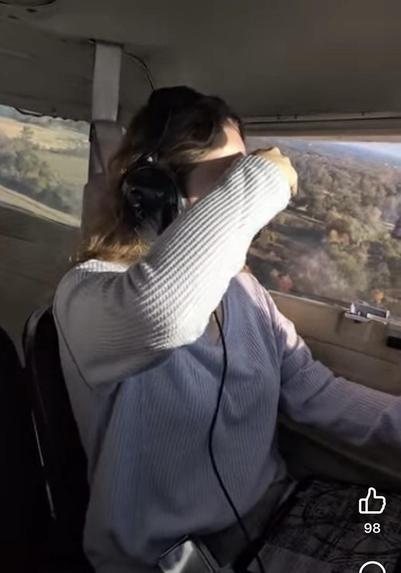 In-cockpit cam still of a young woman with long auburn hair wearing a lavender sweater piloting a plane. Sunlight is streaming in all around, and there are fields and trees on the ground below.