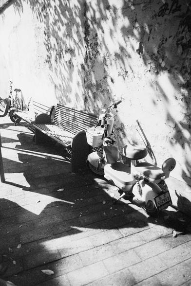 Ilford HP5 Plus 400 (FF)

English Alt Text: A black-and-white photo of a quiet urban sidewalk. Two wooden benches sit against a weathered wall with peeling paint. Beside them are two parked scooters and a bicycle. Dappled sunlight filters through tree leaves above, casting intricate shadows on the wall and pavement. The license plate on the nearest scooter reads “SS2963.” The scene feels still and everyday, evoking a sense of calm in a city setting.

中文替代文字： 这是一张黑白照片，描绘了城市街道的一角。两张木质长椅靠着一面斑驳的墙壁，…