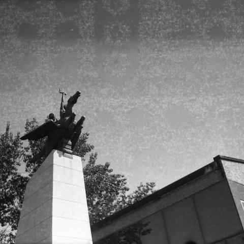 Ilford HP5 Plus 400 (6x6)

English Alt Text: A black-and-white photograph of a tall monument featuring a statue of a winged horse, likely Pegasus, with a human figure riding it. The rider holds a trident-like object aloft, suggesting themes of mythology or heroism. The statue is mounted on a smooth stone pedestal. In the background, trees and a building with a slanted roof are visible. The sky is clear, and the grainy texture of the photo suggests it was taken with film. The composition emphasi…