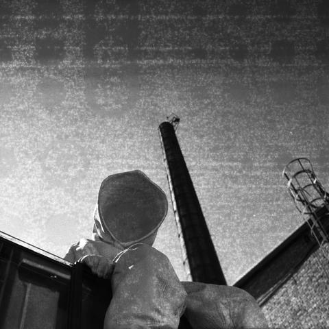 Ilford HP5 Plus 400 (6x6)

English Alt Text: A black and white photo shows a statue of a person wearing a full-body protective suit with a hood, standing on top of  a building by the side of a tall industrial chimney. The perspective is from below, emphasizing the towering height of the chimney. The person’s gear suggests a hazardous or contaminated environment. A metal ladder is attached to the building, reinforcing the industrial setting. The dramatic lighting and monochrome tones evoke a sen…
