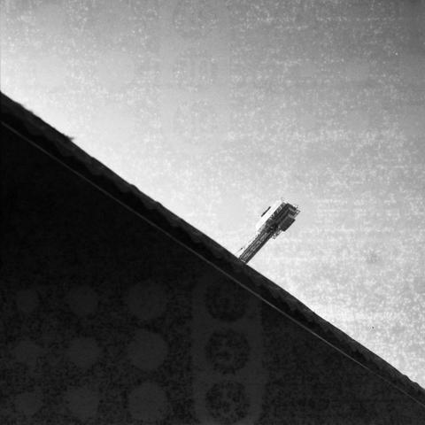 Ilford HP5 (6x6)

English Alt Text: A black-and-white photograph showing a construction crane rising above a sharply angled roofline. The roof cuts diagonally across the image from bottom left to top right, creating a dynamic composition. The crane’s arm extends into the grainy sky, which dominates the upper half of the frame. The contrast between the dark roof and bright sky emphasizes architectural geometry and industrial presence. The image feels stark and modern, with a minimalist aesthetic…