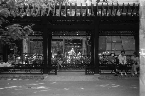 Ilford Pan 100 (FF)

English Alt Text:
A black-and-white photo of a shaded public seating area under a pergola-style structure. Several people sit on benches, some using phones, others chatting. The background includes a glass storefront with reflections and Chinese advertisements, one showing a person. Trees cast dappled light on the ground. The scene captures a moment of urban social life, blending architecture, nature, and human interaction.

中文替代文本：
一张黑白照片，展示一个带棚架的公共座椅区。几位市民坐在长椅上，有人使用手机，有人交…
