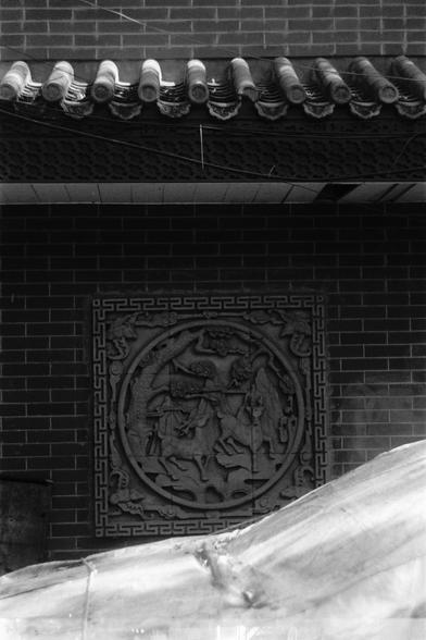 Ilford Pan 100 (FF)
English Alt Text:
A black-and-white image of a traditional Chinese architectural wall. The upper section features a tiled roof with ornate end caps. Beneath the roof is a decorative frieze with repeating patterns. The central focus is a circular stone relief embedded in a brick wall, depicting a detailed scene with human and animal figures, possibly mythological. The relief is framed by a square border with geometric motifs. A translucent plastic sheet partially obscures the…