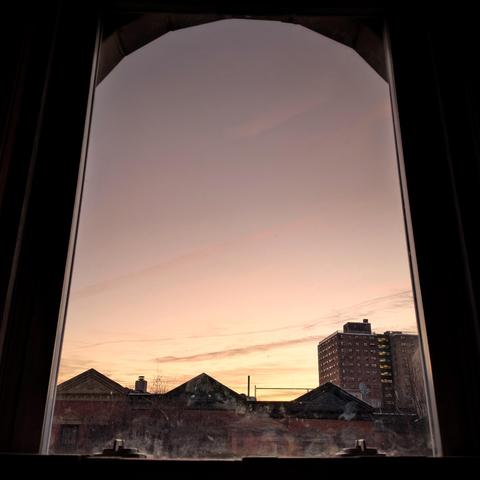Looking through an arched window ten minutes before sunrise the cloudless sky fades from pale yellow at the horizon to rose gray up above. A few small horizontal lines of cloud are above the horizon. Pointed roofs of Harlem brownstones are silhouetted across the street, and a taller apartment building can be seen in the distance. The window is grimy on the bottom.