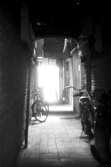 Ilford Pan 100 (FF)

English Alt Text: A black-and-white photo of a narrow alleyway with brick walls on both sides. The alley is paved with tiles and leads to a brightly lit exit in the distance. Several bicycles are parked along the walls, casting shadows on the ground. The lighting creates a dramatic contrast between the dark interior and the bright light at the end, emphasizing depth and texture. The scene feels quiet and urban, with a sense of solitude and anticipation.

中文替代文字：
这是一张黑白照片，展示…