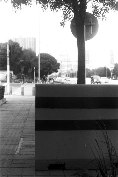 Ilford Pan 100 (FF)

English Alt Text: A black-and-white urban street scene. In the foreground, a large rectangular object with horizontal stripes sits on a brick-paved sidewalk. Behind it stands a tree partially obscuring a circular traffic sign. The background shows a busy street with moving vehicles, pedestrians walking and crossing, and tall buildings lining the road. The photo captures a moment of everyday city life with a mix of natural and man-made elements.

中文替代文字：
这是一张黑白城市街景照片。前景中，一个带…