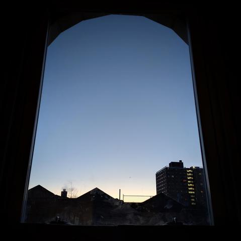 Looking through an arched window twenty-six minutes before sunrise the cloudless sky fades from white at the horizon to baby blue up above. Pointed roofs of Harlem brownstones are silhouetted across the street, and a taller apartment building can be seen in the distance. The window is grimy on the bottom.