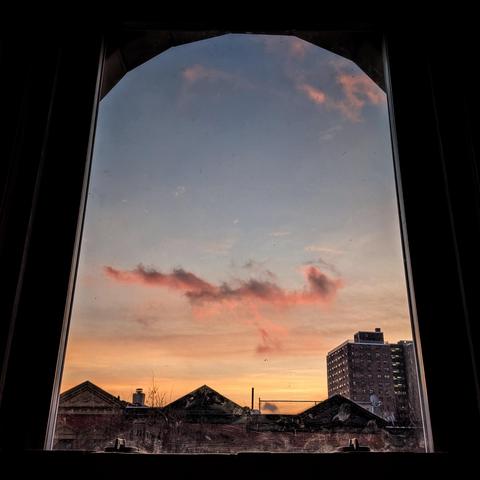 Looking through an arched window two minutes after sunrise the sky fades from orange at the horizon to pale blue up above. Horizontal streaks of orange yellow and gray are above the horizon. A dramatic pink and gray cloud is in the middle of the frame and a few wispy pink clouds are far up above. Pointed roofs of Harlem brownstones are silhouetted across the street, and a taller apartment building can be seen in the distance. The window is grimy on the bottom.