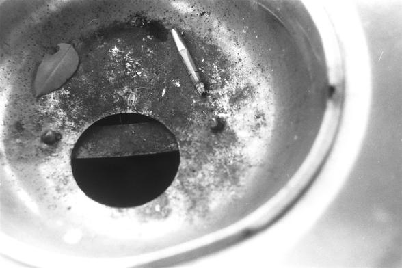 Ilford Pan 100 (FF)

English Alt Text:
A close-up black-and-white image of a metal ashtray. Inside, there’s a partially smoked cigarette, a dry leaf, and scattered ashes. The ashtray has a circular opening in the center and shows signs of frequent use with stains and debris. The composition evokes themes of habit, decay, and stillness, offering a quiet, contemplative moment frozen in time.

中文替代文本：
这是一张黑白特写照片，展示一个金属烟灰缸。烟灰缸内有一根抽了一半的香烟、一片干叶和散落的烟灰。中央有一个圆形开口，表面污渍斑斑，显示出频繁使用的痕迹。画面传达出习惯、衰败与静止的主题，呈现一个凝…