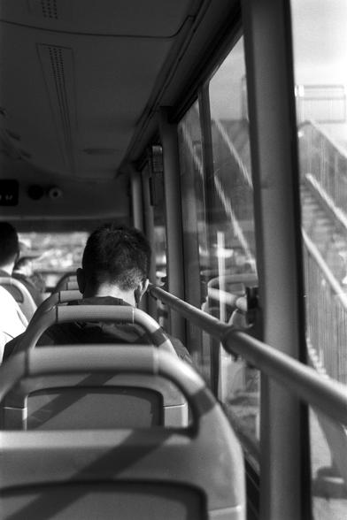 Ilford Pan 100 (FF)

English Alt Text:
A black-and-white photo taken from inside a public bus. The foreground shows the back of a person’s head seated near a window, with another passenger seated further ahead. The bus seats have modern headrests and handles. Sunlight filters through the window, casting reflections of the interior and revealing an exterior view of a staircase and railing, suggesting the bus is near a pedestrian overpass or station. The mood is quiet and observational, capturing…