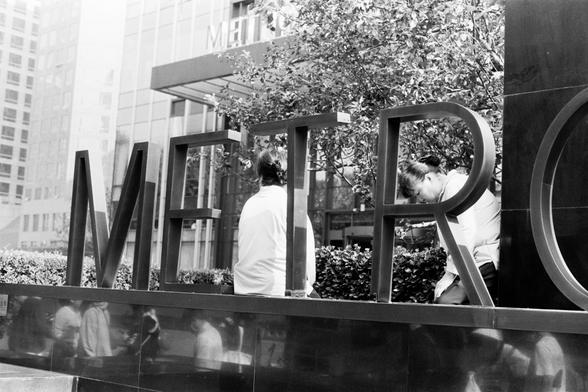 Ilford Pan 100 (FF)

English Alt Text:
A black-and-white urban photograph featuring a large metal sculpture spelling “METRO” in capital letters. The sculpture stands in front of a modern glass building, which also displays the word “METRO” above its entrance. Two people are partially visible behind the letters—one seated, one leaning forward. Reflections of pedestrians appear on the glossy surface below. Trees and bushes add greenery to the scene. The composition blends architecture, typography…