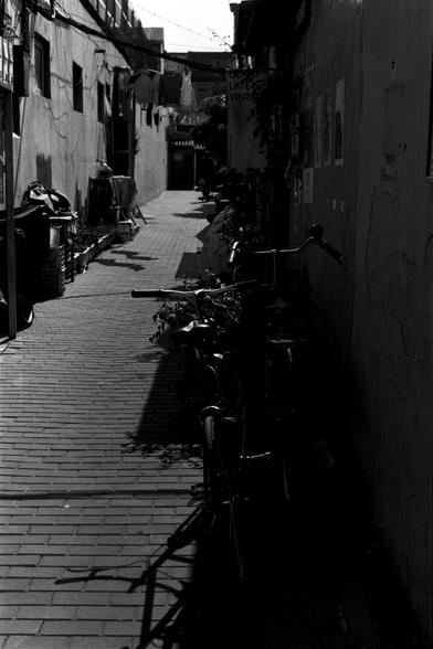 Ilford Pan 100 (FF)

English Alt Text: A narrow brick-paved alleyway in a dense urban setting, captured in black and white. On the left side, laundry hangs from a line above, with containers and potted plants scattered below. The right side features bicycles leaning against a shadowed wall adorned with posters. A sign overhead reads “GOOD LATES VINTA,” possibly part of a vintage shop name. The alley stretches into the distance, ending at a small gate or doorway. The interplay of light and shado…