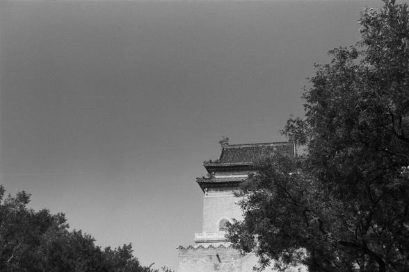 Ilford Pan 100 (FF)

English Alt Text: A black-and-white photo captures the upper part of a traditional East Asian building, partially hidden by trees. The roof has upturned eaves and decorative carvings, with a small sculpture at its peak. The structure appears to be made of stone or brick and includes a circular window. The trees frame the building, adding depth and contrast to the image, which highlights the elegance of historical architecture.

中文替代文字： 这是一张黑白照片，展示了一座传统东亚建筑的上部，部分被树木遮挡。屋顶翘角精致…