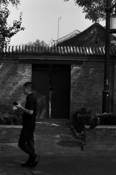 Ilford Pan 100 (FF)

English Alt Text: A black-and-white photo shows a man walking past a traditional Chinese building. The structure has a curved tiled roof and a large double door set into a brick wall. The man wears dark clothing and holds a small object, possibly a phone or wallet. A streetlamp stands nearby, and trimmed bushes line the base of the wall. Trees cast shadows across the scene, creating a peaceful urban moment that blends modern life with cultural heritage.

中文替代文字： 这是一张黑白照片，画面…