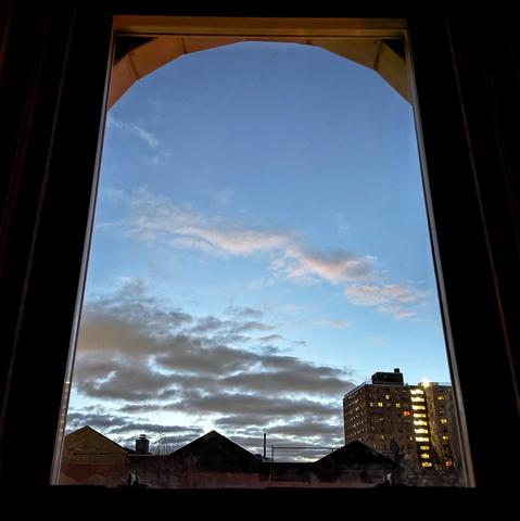 Looking through an arched window thirty nine minutes before sunrise the blue sky is marked with diagonal swaths of gray and white clouds. Pointed roofs of Harlem brownstones are silhouetted across the street, and a taller apartment building can be seen in the distance.