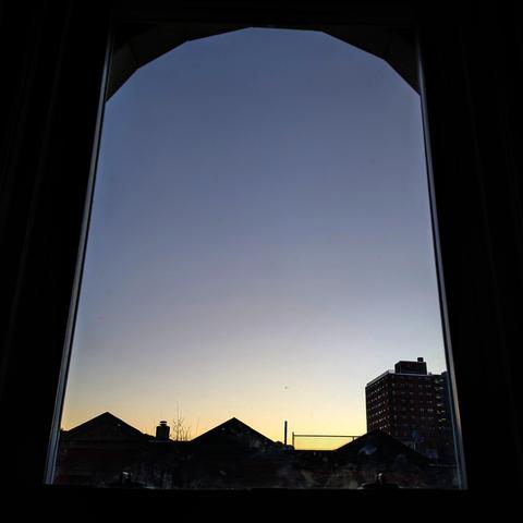 Looking through an arched window three minutes before sunrise the cloudless sky fades from pale lemon yellow at the horizon to slate blue up above. Pointed roofs of Harlem brownstones are silhouetted across the street, and a taller apartment building can be seen in the distance.